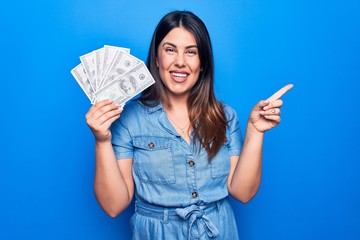 Young beautiful brunette woman holding dollars banknotes over isolated blue background smiling happy pointing with hand and finger to the side