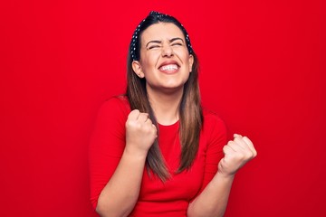 Young beautiful brunette woman wearing casual t-shirt standing over isolated red background celebrating surprised and amazed for success with arms raised and eyes closed
