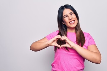 Young beautiful brunette woman wearing casual pink t-shirt standing over white background smiling in love doing heart symbol shape with hands. Romantic concept.