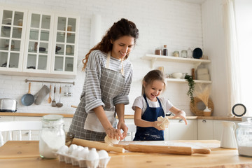 Excited pretty young woman in apron making homemade pastry with cute small child daughter. Smiling little preschool girl playing with dough, enjoying cooking process with happy mother in kitchen.