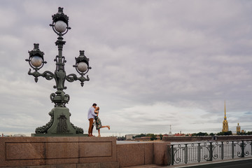 Fototapeta premium Kissing couple wearing casual standing on the bridge close to ancient street lamp in old town. Lovers in the city.
