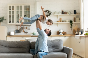 Happy young man lifting small preschool child daughter in air sitting on comfortable sofa in modern...