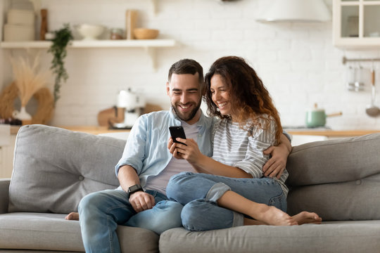 Happy young wife showing funny video in social network on cellphone to laughing husband. Smiling spouses shopping online web surfing, relaxing together on sofa in modern studio kitchen living room.