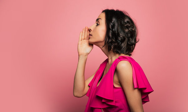 Women's secrets. Close-up photo of a brunette woman in magenta dress, who is standing sideways and holding her hand near her mouth while whispering.