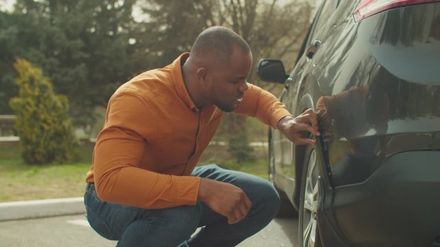 Frustrated worried african male driver checking car scratches and dents after road accident. Unhappy black man looking at damaged scratched vehicle outdoors, being disappointed and upset.