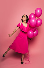 True princess. Full-length photo of overjoyed girl in a fuchsia dress, who is standing on her left leg and holding a bunch of balloons in her left hand.