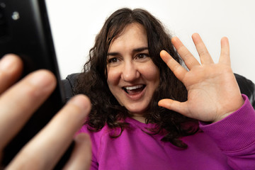 Fototapeta premium Señora blanca de edad media saluda en una videollamada con cara alegre sonriendo y con la mano abierta. Tiene un jersey violeta y un movil negro. Hay un fondo blanco.