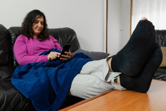 Señora blanca de edad media está sentada en un sofá negro mirando su móvil negro con los pies en una mesa. Tiene un calcetín roto. Está tapada con una manta azul.