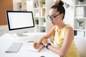 Busy young woman with hair bun sitting at desk with computer and writing down in diary while...