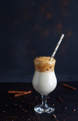 Dalgona coffee, a modern korean smooth, creamy and whisked coffee with milk and ice with cinnamon sticks. Healthy cocktail with soy milk and carob in glass with paper straw on black background. Vertic