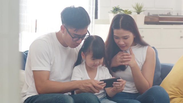 Happy Asian family dad, mom and daughter playing funny game online on smartphone sitting sofa in room at house. Self-isolation, stay at home, social distancing, quarantine for coronavirus prevention.