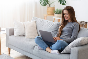 Smiling young lady sitting with laptop on couch at home