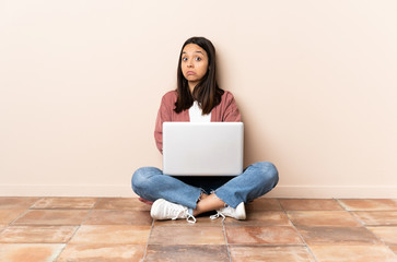 Young mixed race woman with a laptop sitting on the floor making doubts gesture while lifting the shoulders