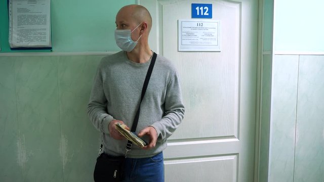 A man in a medical mask stands in a queue in the corridor of the hospital under the door of the office and waits for a doctor's appointment. Turn to the therapist.