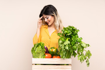 Teenager farmer girl with freshly picked vegetables in a box isolated on beige background laughing