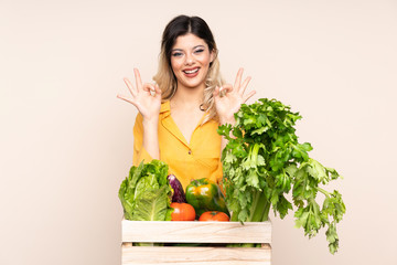 Teenager farmer girl with freshly picked vegetables in a box isolated on beige background showing an ok sign with fingers