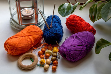 Home hobbies. Composition in bright colors of yarn, beads, pins and knitting needles. Nearby is a candle in a metal candlestick and green leaves of a flower. What to do at home.