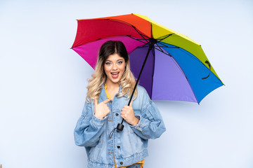 Teenager russian girl holding an umbrella isolated on blue background with surprise facial expression