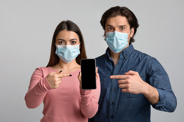 Shocked Couple In Medical Masks Pointing At Smartphone With Black Screen