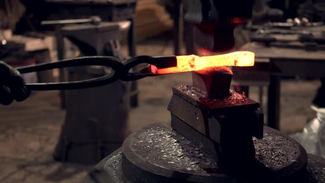Blacksmith holding blacksmith tongs with red hot iron bar working at large industrial hammer