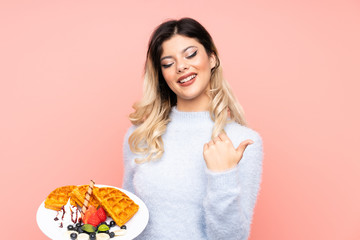 Teenager girl holding waffles on isolated pink background pointing to the side to present a product