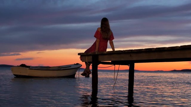 Young slender girl with long blonde hair, in red dress with bare shoulders, sits with her back on a wooden pier or bridge and looks at a beautiful sunset and a wooden boat on water. Relax, loneliness.