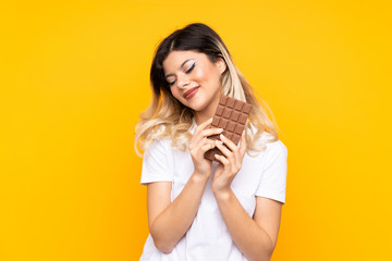 Teenager girl isolated on  yellow background taking a chocolate tablet and happy