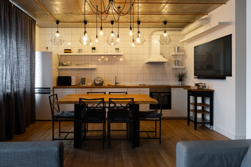 Modern scandinavian an eat-in kitchen interior design with big wooden table and chairs against light wood floor, bright white walls and furnitures with TV, appliances and hanging light bulbs