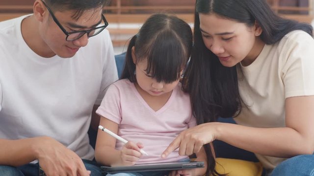 Happy Asian family dad, mom and daughter using computer tablet technology sitting sofa in living room at house. Self-isolation, stay at home, social distancing, quarantine for coronavirus prevention.