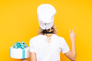 Teenager girl pastry holding a big cake isolated on yellow background pointing back with the index finger