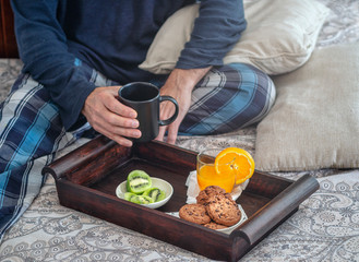 Healthy breakfast on a wooden tray in bed Man holding a cup. Glass of orange juice, kiwi and cookies. Brown green and orange. White cushions.