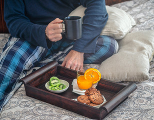 Healthy breakfast on a wooden tray in bed Man holding a cup. Glass of orange juice, kiwi and cookies. Brown green and orange. White cushions.