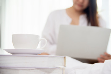 Close up on coffee cup in morning with blurred  woman sitting on bed working on laptop computer  in bedroom at home