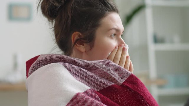 Sick caucasian woman wrapped into blanket covering her mouth with napkin and coughing, then removing medical thermometer and checking temperature while having cold and staying at home