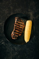 A single sirloin cut steak that has been grilled outdoors on a gas grill with a fork and knife and a piece of corn on the cob