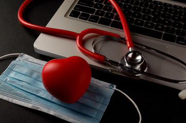 Stethoscope, face mask and red heart with laptop on dark background. Health concept. Selective focus.