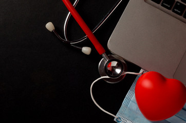 Stethoscope, face mask and red heart with laptop on dark background. Health concept. Selective...