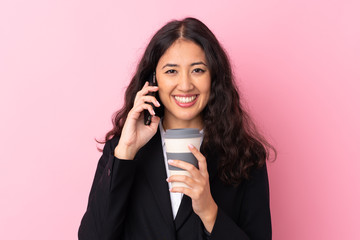 Mixed race business woman holding coffee to take away and talking to mobile over isolated pink background