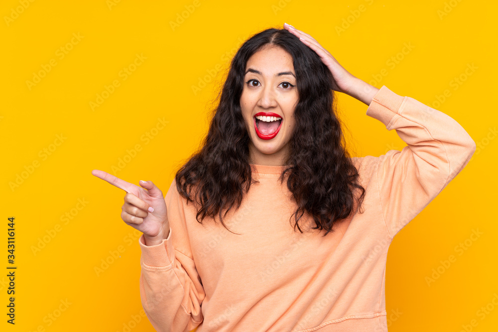 Wall mural spanish chinese woman over isolated yellow background surprised and pointing finger to the side