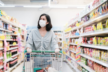 Asian woman wearing face mask and rubber glove push shopping cart in suppermarket departmentstore. Girl choosing, looking grocery things to buy at shelf during coronavirus crisis or covid19 outbreak.