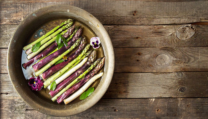 Set of raw white, green, purple asparagus in copper basin with violet and mint. Old wooden rustic background. Organic healthy eating concept. Copy space. Overhead.