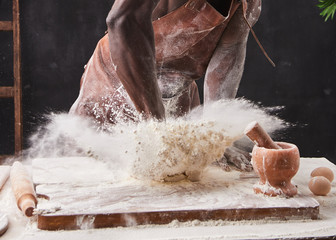 Dark skin baker man kneads Dough in the kitchen. White flour flies in air on black background, pastry chef claps hands and prepares yeast dough for pizza pasta.