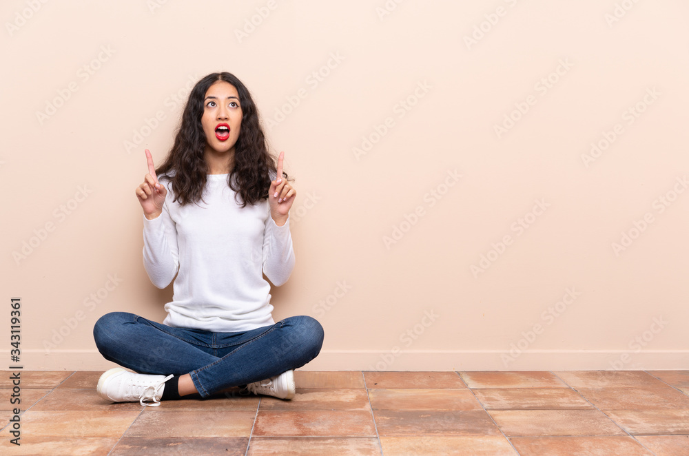 Wall mural Young woman sitting on the floor surprised and pointing up