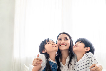 Obraz na płótnie Canvas Asian family mother, son and daughter sit on white bed with happiness and smile in bedroom. Mom hug boy and girl playing together feeling fun. People look above. House property mortgage advertisement.