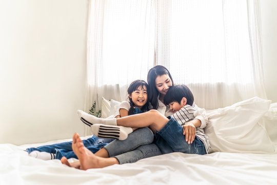Asian family mother, son and daughter sit on white bed with happiness and smile in bedroom. Young boy and girl hug single mom smiling enjoy playing together at home. Love and warm touch from parent.