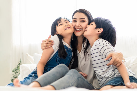 Asian Mother, Son And Daughter Sit On White Bed With Happiness And Smile In Bedroom. Mom Hug Boy And Girl Playing Together. Children Kissing Mom With Love In Warm House. Happy Family Relationship.