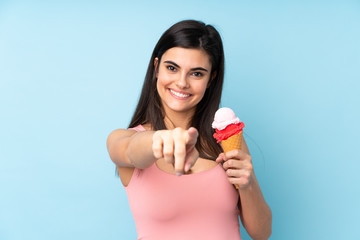 Young woman holding a cornet ice cream over isolated blue background points finger at you with a confident expression