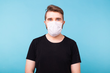 Handsome young man in black t-shirt wearing medical mask on isolated blue background. Covid-19, coronavirus and virus concept.