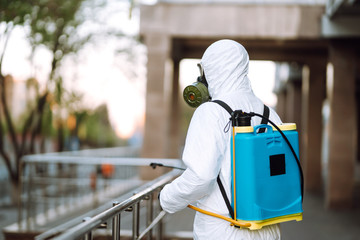 Man in protective suit  and mask sprays disinfector onto the railing in the empty public place at dawn in the city of quarantine. Covid -19. Cleaning concept.