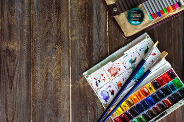 art workplace, color pencils, brushes and watercolor paints, copy space. Flat lay on wooden table
